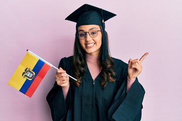Young hispanic woman wearing graduation uniform holding ecuador flag smiling happy pointing with hand and finger to the side