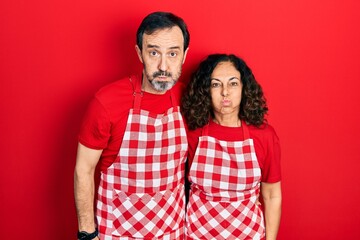 Middle age couple of hispanic woman and man wearing cook apron puffing cheeks with funny face. mouth inflated with air, crazy expression.