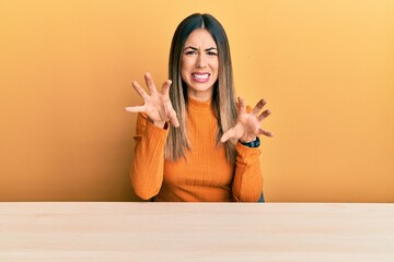 Young hispanic woman wearing casual clothes sitting on the table smiling funny doing claw gesture...