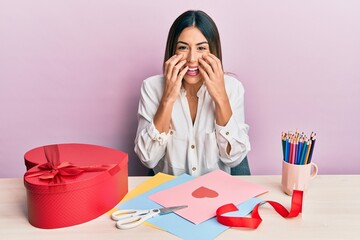 Young hispanic woman making valentine gift sitting on the table laughing and embarrassed giggle...
