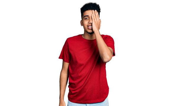 Young african american man wearing casual red t shirt covering one eye with hand, confident smile on face and surprise emotion.