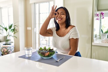 Obraz na płótnie Canvas Young hispanic woman eating healthy salad at home doing ok gesture with hand smiling, eye looking through fingers with happy face.