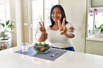 Obraz na płótnie Canvas Young hispanic woman eating healthy salad at home smiling looking to the camera showing fingers doing victory sign. number two.