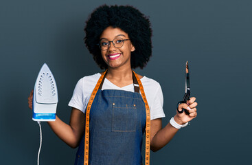 Young african american woman dressmaker designer wearing atelier apron holding iron and scissors...