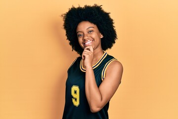 Fototapeta na wymiar Young african american woman wearing basketball uniform looking confident at the camera with smile with crossed arms and hand raised on chin. thinking positive.