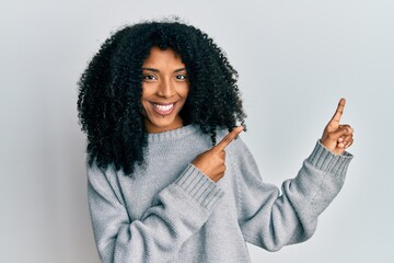 African american woman with afro hair wearing casual winter sweater smiling and looking at the camera pointing with two hands and fingers to the side.