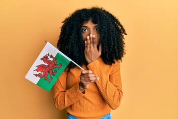 African american woman with afro hair holding wales flag covering mouth with hand, shocked and afraid for mistake. surprised expression