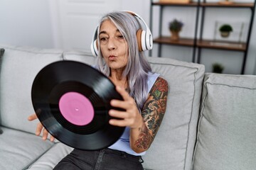 Middle age grey-haired woman listening to music holding vinyl disc at home looking at the camera blowing a kiss being lovely and sexy. love expression.