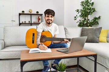 Young hispanic man playing classical guitar and using laptop at home.