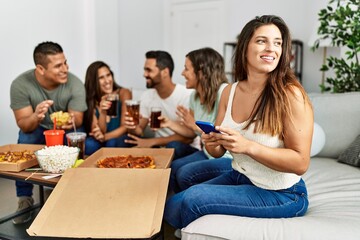 Group of young hispanic friends eating italian pizza sitting on the sofa. Woman smiling happy and using smartphone at home.