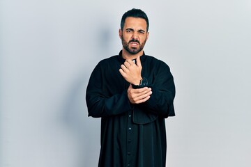 Handsome hispanic man with beard wearing catholic priest robe suffering pain on hands and fingers,...