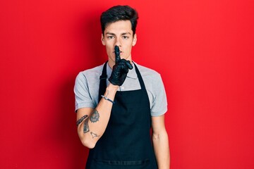 Young hispanic man wearing apron asking to be quiet with finger on lips. silence and secret concept.