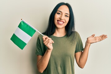 Young hispanic girl holding nigeria flag celebrating achievement with happy smile and winner...