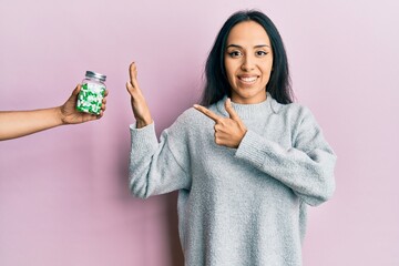 Young hispanic girl rejecting jar of pills with hand smiling happy pointing with hand and finger