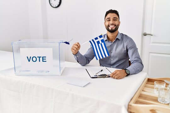 Young handsome man with beard at political campaign election holding greece flag looking positive and happy standing and smiling with a confident smile showing teeth