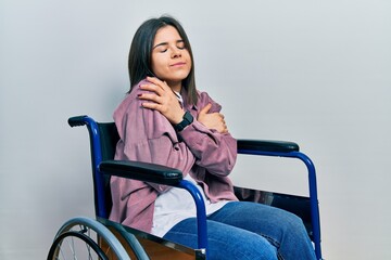 Young brunette woman sitting on wheelchair hugging oneself happy and positive, smiling confident....