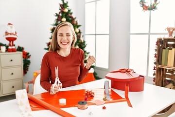 Caucasian young blonde woman sitting on the table doing christmas handcrafts screaming proud, celebrating victory and success very excited with raised arms