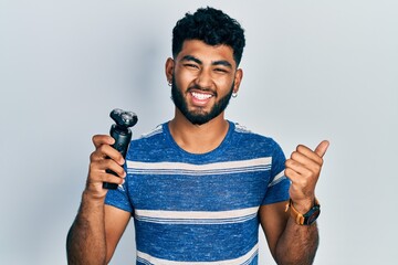 Arab man with beard holding electric razor machine screaming proud, celebrating victory and success...