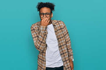 Young african american man with beard wearing casual clothes and glasses smelling something stinky and disgusting, intolerable smell, holding breath with fingers on nose. bad smell