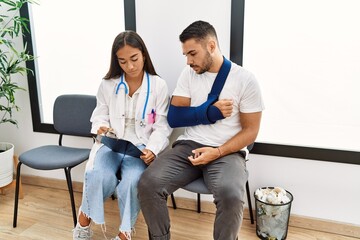 Youg latin injuried man wearing sling talking with the doctor at clinic waiting room.