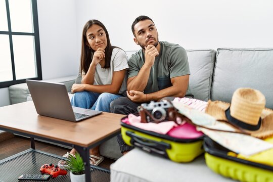 Young interracial couple packing summer clothes in suitcase looking at laptop serious face thinking about question with hand on chin, thoughtful about confusing idea
