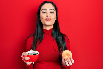 Young hispanic girl drinking coffee and eating pastry looking at the camera blowing a kiss being...