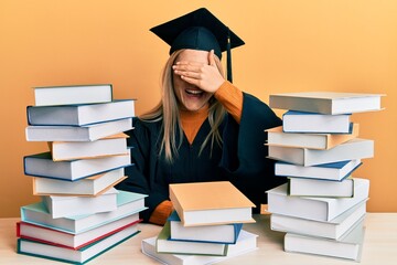 Young caucasian woman wearing graduation ceremony robe sitting on the table smiling and laughing with hand on face covering eyes for surprise. blind concept.