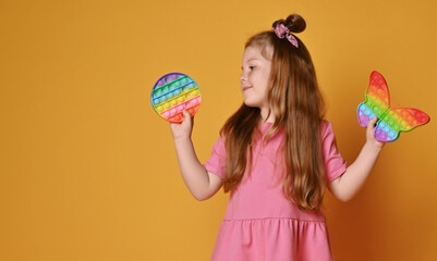 Happy smiling red-haired kid girl in pink clothes holds, demonstrates two new sensory rainbow color...