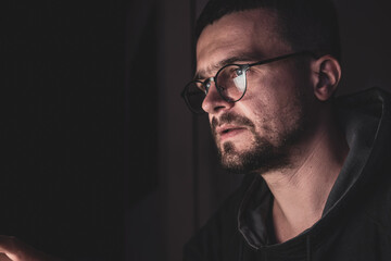 A man with glasses in the dark looks at the computer screen, copy space.