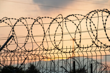 Metal fence with barbed wire on the border, against the background of a pink sunset.