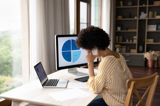 Concentrated young african ethnicity businesswoman employee worker holding phone call conversation with client, working with paper documents, analyzing economic data on computer, multitasking.
