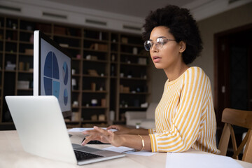 Focused millennial african ethnicity businesswoman in eyeglasses working on computer, using monitor...