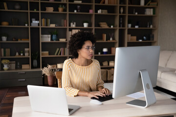 Concentrated young mixed race businesswoman entrepreneur employee in eyeglasses looking at computer...