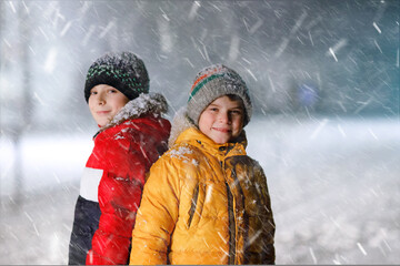Two little kids boys of elementary class walking to school during snowfall on early dark morning. Happy children having fun and playing with first snow. Siblings ans friends in colorful winter clothes