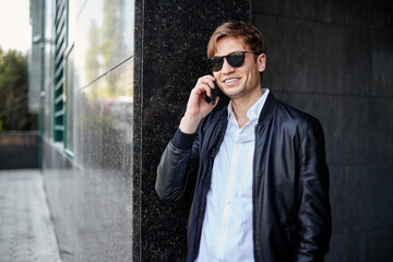 Portrait of a handsome young businessman in black sunglasses and leather jacket, talking on a cell phone, outside.