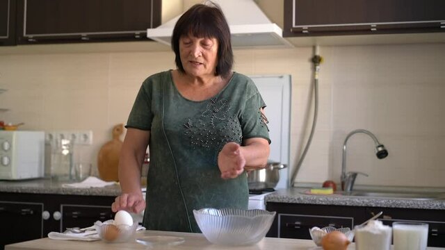 Happy elderly woman uses fresh organic food, flour milk eggs, kneading dough for baking, cookies with her hands at home in the kitchen. Mature old lady makes homemade food