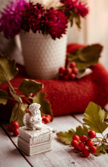 Ceramic white figure of an angel reading a book against a background of warm knitted things and autumn flowers in a white mug. Cozy homely autumn composition on a light wooden background