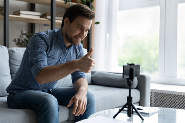 Smiling young man showing thumb up, using smartphone on tripod, satisfied blogger influencer recommending service, recording video for social network on mobile device, sitting on couch at home