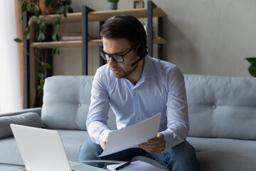 Confident businessman in wireless headset and glasses talking, working, consulting client online about deal terms, holding documents, serious man engaged in internet meeting, conference at home