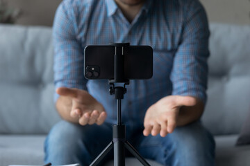 Crop close up man recording video, using smartphone on tripod, popular blogger shooting vlog for social network, sitting on couch at home, mentor coach speaking leading webinar, broadcasting live