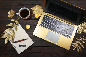 golden laptop computer, gold painted autumn leaves, coffee cup and a notepad with pen on a dark...