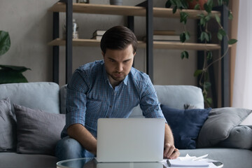 Serious man using laptop, working or studying online, sitting on couch at home, focused confident businessman freelancer or student looking at computer screen, watching webinar, taking notes