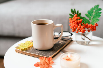 Still life book, candle, rowan berry and a cup of tea or coffee in the living room on a table, home decor in a cozy house. Autumn weekend concept, blanket and plaid. Fallen leaves and home decoration