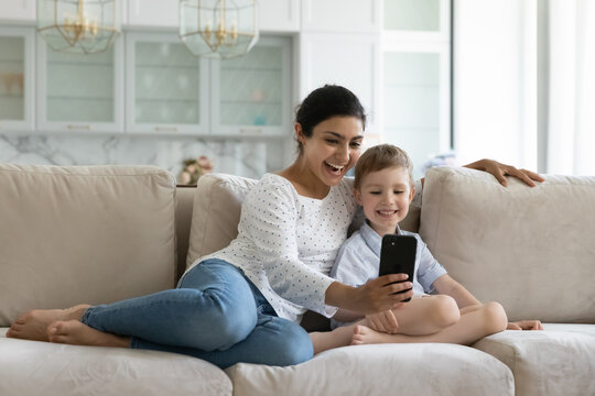 Happy bonding young indian mother and little adorable caucasian kid son using cellphone, streaming funny stories in social networks, posing for selfie photo, having fun together at home. - Powered by Adobe