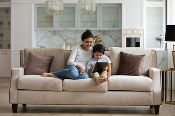 Happy young indian mum or nanny using digital computer tablet with cute little kid boy, resting together on comfortable sofa, playing online games, shopping in internet store or web surfing.
