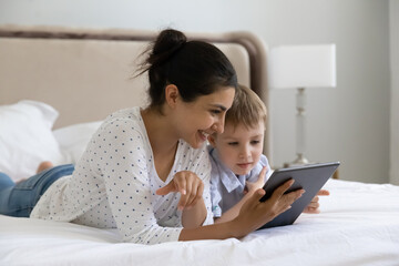 Caring smiling young indian woman using digital computer tablet applications with happy small preschool kid son, playing games resting on bed, parental control, tech addiction concept.
