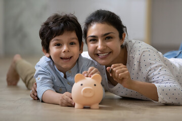 Portrait of happy indian ethnic family lying on floor with small piggybank. Caring young asian mum...