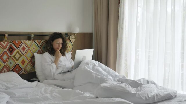 Happy curly haired young woman in white bathrobe holds laptop thinks types and smiles sitting on hotel bed with colored headboard