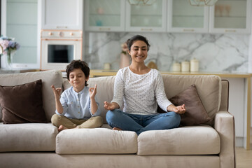 Happy small child boy sitting in lotus position on comfortable couch with joyful asian indian mother, practicing together yoga breathing exercise, doing asanas, enjoying domestic healthcare activity.