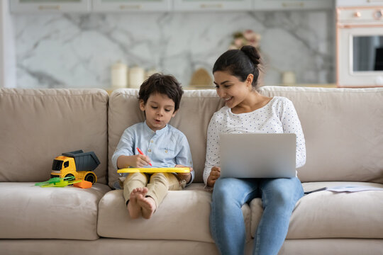 Smiling young indian asian woman taking break pause time working distantly on computer, playing with adorable small child son, sitting together on comfortable couch, parenting and career concept.